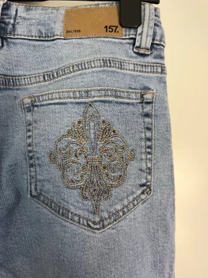 Ljusblå  jeans från lager 157 - Säljer ett par ljusblå jeans från 157 med snyggt broderi på bakfickan. Jeansen har en lätt bootcut och klassisk femficksmodell. Tillverkade i denim med cool vintagekänsla och detaljer i guldtråd på fickan. Perfekta för en trendig och avslappnad stil. Aldrig anvönd då de va försmå