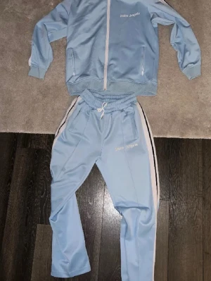 Ljusblått loungewear-set Palm Angels - Snyggt ljusblått loungewear-set från Palm Angels med vit dragkedja och vita sidoremsor. Setet består av en zip-jacka med fickor och matchande byxor med resår i midjan och dragsko. Båda delarna har Palm Angels-logga broderad på bröst och lår. Qr kod finns