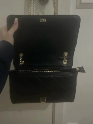 Svart quiltad handväska Valentino - Snygg svart quiltad handväska från Valentino med guldfärgade kedjehandtag och detaljer. Väskan har ett klassiskt rektangulärt format, dragkedja och tryckknapp. Materialet ser ut att vara konstläder. Perfekt för dig som gillar stilrena accessoarer.