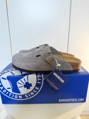 Birkenstock Boston tofflor grå mocka - Säljer ett par Birkenstock Boston tofflor i mörkgrå mocka med justerbart spänne i metall. Tofflorna har en ergonomisk korksula och en mjuk ovandel som ger en skön passform. Perfekta för dig som vill ha både stil och komfort.
