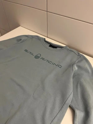 Ljusblå Sail Racing sweatshirt - Ljusblå sweatshirt från Sail Racing med rund halsringning och tryckt logga på bröstet. Tröjan har en enkel och stilren design med långa ärmar och mjukt material, perfekt för en avslappnad look.