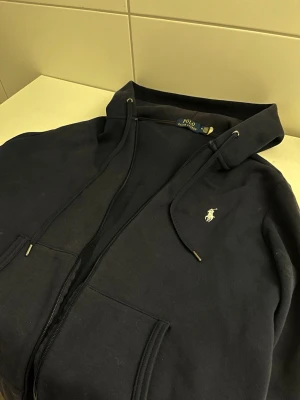 Mörkblå hoodie från Polo Ralph Lauren - Mörkblå hoodie med dragkedja från Polo Ralph Lauren. Klassisk design med huva och snörning, samt den ikoniska broderade loggan på bröstet. Mjuk bomullskvalitet och kängurufickor framtill. Perfekt för en avslappnad och stilren look.