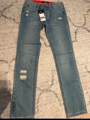 Blå slitna jeans från dB, raka ben - Snygga blå jeans från dB med raka ben och slitna detaljer framtill. Klassisk femficksmodell med röd logga på bakfickan och normalhög midja. Jeansen har en cool, avslappnad look och är gjorda i denim med lätt stretch för extra komfort.