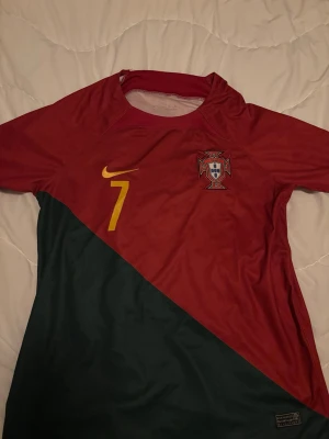 Portugal Nike fotbollströja röd/grön - Portugal fotbollströja från Nike med röd överdel och mörkgrön diagonal nederdel. Nummer 7 i gult på bröstet och Portugals landslagsemblem. Tillverkad i lätt och ventilerande material, perfekt för träning eller match.