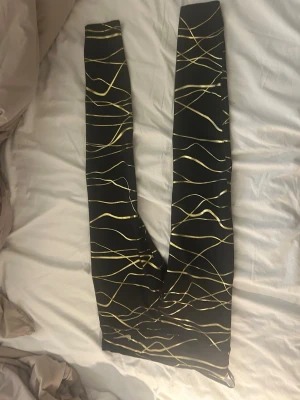 Nike tights med guldmönster - Svarta Nike tights med glansiga, abstrakta guldlinjer över hela tyget. Tight passform och stretchigt material som sitter bekvämt. Finare i verkligheten än på bilden. Storlek S/M (mindre M) så gott som nya!