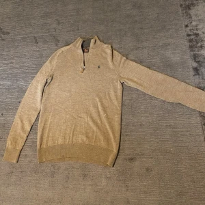 Beige halvzip-tröja från Morris - Snygg guld/beige stickad tröja från Morris med halvzip och liten broderad logga på bröstet. Tröjan har långa ärmar, ribbade muddar och krage. Perfekt för en stilig och avslappnad ”Old Money” look.