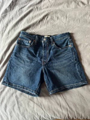 Blå jeansshorts från Levi's - Klassiska blå jeansshorts från Levi's med fem fickor och gylf med knapp. De har en rak passform och tydliga sömmar, samt Levi's ikoniska läderpatch bak i midjan. Perfekta för en avslappnad stil under varmare dagar.