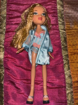 Vintage Bratz docka – underkläder & morgonrock – tidigt 2000-tal - Original Bratz-docka från den tidiga Y2K-eran med ljusblont hår och klassisk Bratz-sminkning. Dockan säljs med matchande underklädesset, satinmorgonrock och klackskor enligt bilderna.  Håret har fina crimpade slingor och typisk tidig 2000-tals-styling som är extra eftertraktad bland samlare. Ansikte, hår och kropp är i mycket fint vintage-skick utan större slitage.  En ovanligare och mer “sleepover/lounge”-inspirerad Bratz-look som verkligen fångar den klassiska Y2K-estetiken.  