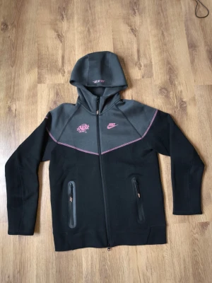 Nytt Nike Tech Fleece "Life of Syn" Limited Edition Set - Storlek L - Säljer ett helt nytt och oanvänt Nike Tech Fleece-set i den exklusiva "Life of Syn"-utgåvan. Dräkten är i nyskick med alla etiketter kvar (10/10). Detta är en unik modell som kombinerar Nikes premiumkomfort med en snygg streetwear-design. Svart bas med kontrasterande rosa sömmar och grafitti-tryck som verkligen sticker ut. Mått (Storlek L): Huvtröja: Längd 62 cm, axelbredd 35 cm. Byxor: Längd 98 cm. Material: Nike Tech Fleece (lätt och värmeisolerande). Skick: Helt ny med tags.