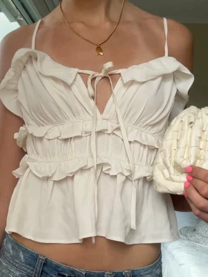 Vit volangblus  - Supersöt vit blus med volanger, smala axelband och knyt framtill. Luftig modell med omlottkänsla och romantiska detaljer. Perfekt till jeans eller kjol för en trendig look. Aldrig använd