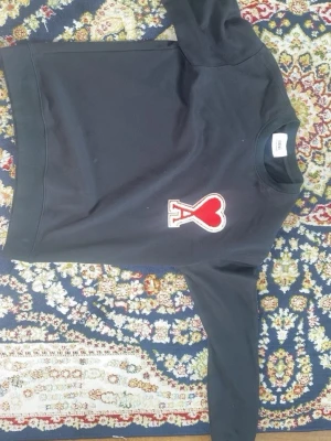 Svart sweatshirt Ami Paris  - Svart sweatshirt från Ami Paris med rund hals och långa ärmar. På bröstet sitter ett stort rött hjärta med bokstaven A broderat. Tröjan har ribbade muddar och är gjord i mjuk bomull för en clean och trendig vibe.
