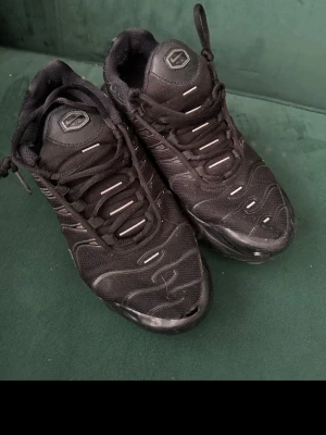 Svarta Nike Air Max Plus sneakers - Säljer ett par svarta Nike Air Max Plus sneakers med klassisk mesh- och syntetöverdel, tydliga vågmönster och snörning. Sulan har gula och röda detaljer undertill och den ikoniska TN-loggan på hälen. Perfekta för dig som gillar streetwear och sportig stil.