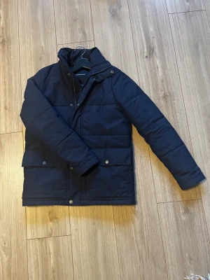 Peak performance jacka  - Säljer en mörkblå pufferjacka med hög krage och två stora fickor med tryckknappar framtill. Jackan har quiltad design och långa ärmar, perfekt för kalla vinterdagar. Materialet är syntet och jackan har en stilren look som passar dig som vill ha något både varmt och snyggt.