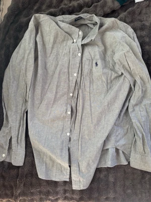 Grå skjorta från Polo Ralph Lauren - Klassisk grå skjorta från Polo Ralph Lauren i 100% bomull. Skjortan har knappar framtill, bröstficka med broderad logga och lång ärm. 