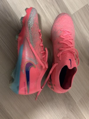 Nike rosa Mercurial fotbollsskor - Säljer ett par Nike Mercurial fotbollsskor i en riktigt snygg rosa färg med silverdetaljer och blått swoosh-märke på sidan. Skorna har snörning, låg profil och en lätt ovandel i syntetmaterial för bra bollkontroll. Sulan är delvis blå och designad för bra grepp på planen.