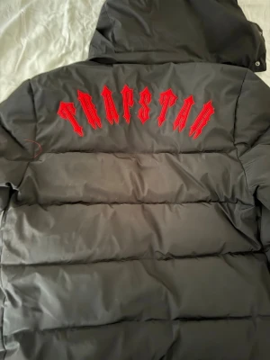 Svart pufferjacka från Trapstar - Säljer en svart pufferjacka från Trapstar med stor röd logga i gotisk stil på ryggen och rund patch på bröstet. Jackan har huva med dragsko och grov dragkedja framtill. Perfekt för dig som vill ha en streetwear-look i vinter.