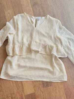 Cremefärgad  blus med volang och struktur - Söt cremefärgad blus från Lindex med diskret strukturmönster och volangdetalj. Blusen har v-ringning, knapp i nacken och långa ärmar i ett lätt, transparent tyg. Perfekt för en trendig och feminin look. Oanvänd 