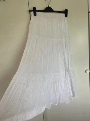 Vit lång asymmetrisk kjol Gina Tricot - Säljer en vit lång kjol med asymmetrisk nederkant och volangdetaljer. Kjolen är gjord i ett tunt, luftigt tyg som ger en bohemisk känsla. Perfekt för sommardagar och festivaler. Ingen synlig logga eller märke.