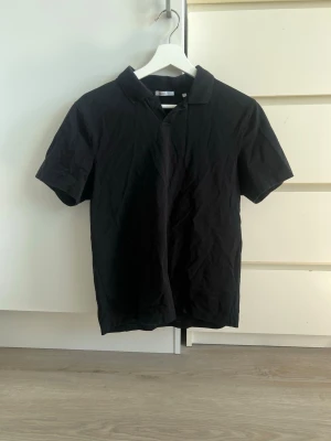 Svart pikétröja från Bläck - Sitter åt det mer slim hållet. Klassisk svart pikétröja från Bläck med korta ärmar och krage. Tillverkad i ett mjukt och bekvämt material som passar perfekt till många olika stilar. Enkel design utan tryck eller mönster, vilket gör den lätt att matcha.