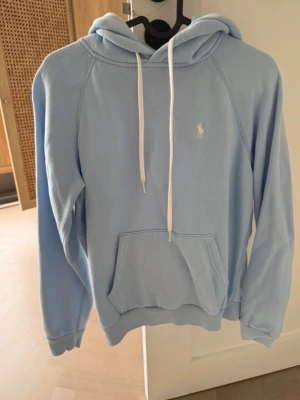Ljusblå hoodie från Ralph Lauren - Snygg ljusblå hoodie från Ralph Lauren med vit broderad logga på bröstet. Hoodien har huva med vita snören och en stor magficka framtill. Mjuk och skön bomullskänsla, perfekt för chill dagar eller när du vill vara extra bekväm.