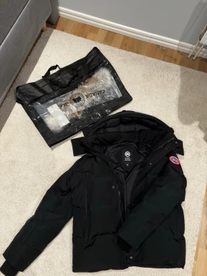 Svart dunjacka från Canada Goose - Säljer en svart dunjacka från Canada Goose med avtagbar pälskant till huvan. Jackan har klassisk patch på ärmen, dragkedja och knappar framtill samt flera fickor. Perfekt för kalla vinterdagar och riktigt varm.