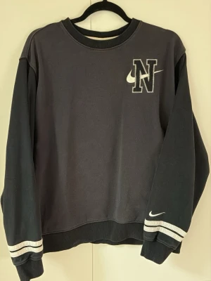 Nike sweatshirt - Svart sweatshirt från Nike i vintage stil. Tröjan har långa ärmar, ribbade muddar och vita ränder vid ärmsluten. Perfekt för en avslappnad streetstyle-look.