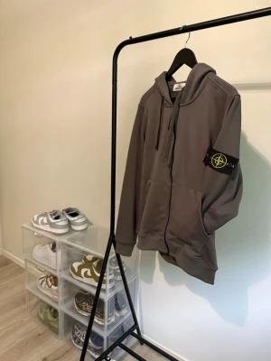 Grå zip hoodie från Stone Island - Säljer en grå zip hoodie från Stone Island med klassisk patch på vänster ärm. Hoodien har dragkedja framtill, två stora fickor och justerbar huva med snören. Tillverkad i mjukt material som känns skönt mot huden. Perfekt för en avslappnad streetwear-look.
