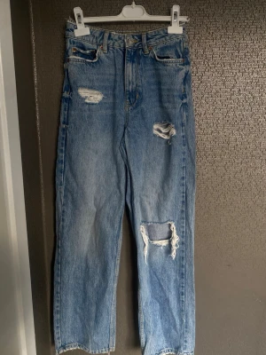 Blå mom jeans med slitningar - Säljer ett par ljusblå mom jeans med flera slitningar och hål på framsidan. Jeansen har hög midja, raka ben och klassisk femficksmodell. Materialet är denim i 100% bomull och de har en avslappnad passform som är trendig och bekväm.