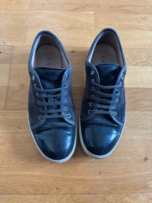Svarta sneakers från Lanvin - Stilrena svarta sneakers från Lanvin med mocka på ovandelen och lackad tå. Skorna har vita sulor och svarta skosnören. Perfekta för dig som gillar en clean och lyxig look. Klassisk låg modell som passar till det mesta.