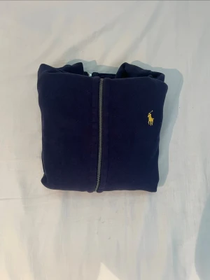 Mörkblå hoodie från Polo Ralph Lauren - Snygg mörkblå hoodie från Polo Ralph Lauren med dragkedja framtill och gul broderad logga på bröstet. Hoodien har vita snören i huvan och är gjord i mjukt bomullsmaterial. Perfekt för en chill och stilren look.