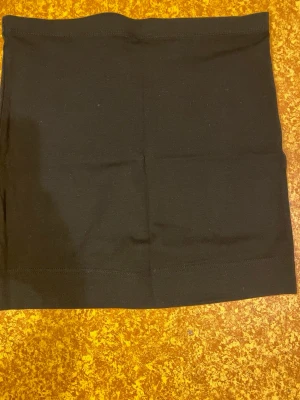 Svart kortkjol från H&M - En enkel och stilren svart kortkjol från H&M i stretchigt bomullsmaterial. Kjolen har en rak passform och är perfekt att matcha med olika toppar för en clean look. Klassisk midja utan detaljer eller mönster.