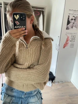 Beige stickad tröja med dragkedja - Mysig beige stickad tröja med halv dragkedja framtill. Tröjan har en grovstickad struktur, långa ärmar och ribbade muddar. Perfekt för lager på lager och en avslappnad stil.