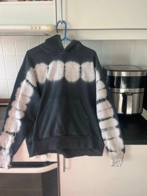 Svart tie-dye hoodie från Weekday - Säljer en svart hoodie från Weekday med vit tie-dye effekt över bröstet och längs ärmarna. Hoodien har en stor känguruficka framtill och klassisk huva. Tillverkad i mjukt bomullsmaterial, perfekt för en avslappnad och trendig look.
