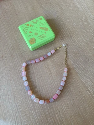 Pastellfärgat pärlhalsband från Anni Lu - Säljer ett halsband från Anni Lu med fyrkantiga pärlor i pastellfärger som rosa, lila och vitt. Halsbandet har guldfärgad kedja och lås. Kommer i original grön ask med guldmönster. Perfekt för dig som gillar färgglada accessoarer.