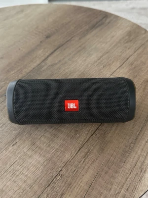 JBL svart bärbar högtalare - Snygg svart bärbar högtalare från JBL med cylinderformad design och robust tygmaterial runt om. Högtalaren har en tydlig JBL-logga i rött och svart samt en praktisk snodd för enkel transport. Perfekt för att ta med sig musiken överallt.