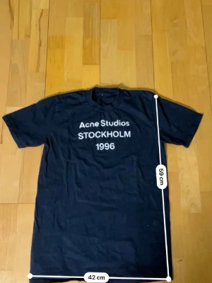 Svart Acne Studios t-shirt - Snygg svart t-shirt från Acne Studios med vit text 'STOCKHOLM 1996' på bröstet. Klassisk rund hals och korta ärmar. Perfekt till jeans eller shorts för en clean och stilren look.