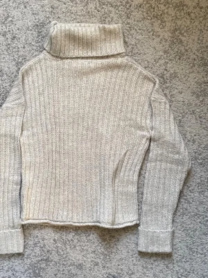 Beige ribbstickad polotröja - Säljer en beige ribbstickad polotröja med hög krage och långa ärmar. Tröjan har en rak passform och breda muddar vid ärmsluten. Perfekt för lager på lager och enkel att matcha med olika outfits.