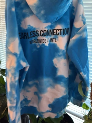 Blåvit hoodie med moln och text - Säljer en blåvit hoodie med molnliknande tie-dye mönster och texten 'FEARLESS CONNECTION WORLDWIDE / INTL' tryckt på både bröstet och ryggen. Hoodien har huva och en stor magficka framtill. Perfekt för dig som gillar streetwear och unika plagg.