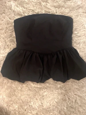 Svart volangig tubtopp - Säljer en svart axelbandslös tubtopp med volang nedtill. Toppen har en bandeauformad överdel och smockad rygg för extra komfort. Perfekt för dig som vill ha en trendig och stilren look.