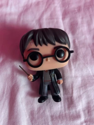 Harry Potter Funko Pop figur - Säljer en Harry Potter Funko Pop figur med svarta glasögon, svart mantel och trollstav. Figuren har detaljer som ärr i pannan och Hogwarts-uniform. Perfekt för samlare eller som dekoration. Originella förpakningen ingår inte 