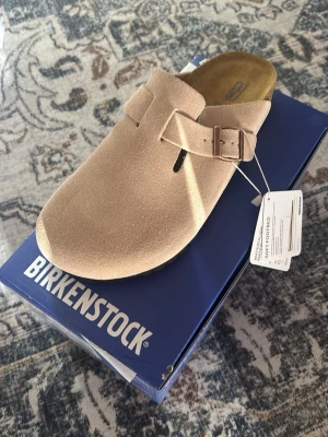 Birkenstock Boston bs ’sand’ - Säljer ett par Birkenstock Boston tofflor i sandfärgad mocka med justerbart spänne. Klassisk slip-in modell med mjuk fotbädd och robust sula. Perfekta för chill dagar hemma eller när du vill ha en clean och avslappnad stil.