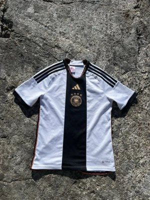 Tysklands fotbollströja Adidas - Säljer en officiell Tyskland fotbollströja från Adidas. Tröjan är vit med en bred svart mittpanel, guldiga detaljer och fyra stjärnor ovanför landslagets emblem. Klassiska Adidas-ränder på axlarna och korta ärmar. Perfekt för match eller träning. Det står 15-16 år men passar dig som brukar ha storlek S