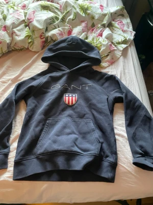Mörkblå hoodie från GANT med logga - Snygg mörkblå hoodie från GANT med broderad logga och amerikanskt sköldmärke på bröstet. Klassisk känguruficka framtill och huva. Perfekt för en avslappnad och stilren look. Mjuk bomullsblandning som känns skön mot huden.