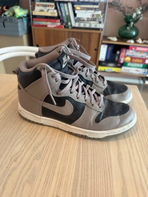 Nike Dunk High grå och svarta sneakers - Nike Dunk High sneakers i grått och svart med klassisk swoosh på sidan. Skorna har högt skaft, perforerad tåbox och matchande grå snörning. Ovandelen är i läder och textil, och sulan är vit med grå detaljer. Perfekta för dig som gillar streetwear och retrostil.