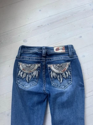 Miss me jeans - Säljer ett par blå Miss Me jeans med snygga broderade detaljer på bakfickorna i form av mönster och fjädrar. Jeansen är mid waist i storlek w24 som motsvarar ungefär xxs🥰 