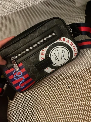 Svart magväska med GG-mönster - Snygg svart magväska med GG-mönster, rödblå randig axelrem och coolt ormmotiv. Väskan har en stor rund patch med text och dragkedja framtill. Materialet är canvas med läderdetaljer och väskan har en modern, rektangulär form.