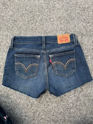 Vintage Levi's jeansshorts med spetsdetaljer - Säljer ett par klassiska blå lågmidjade jeansshorts från Levi's med råa kanter och snygga detaljer på fickorna!!😍Shortsen har fem fickor och bälteshällor. De har spetskant på fickorna, små pärlor och en stjärna framtill! Perfekta för sommaren och riktigt sköna att bära💞Så coola och gulliga, ger din outfit extra snygghet! Sydda och dekorerade av mig med noggrannhet, passion & kärlek❤️Hör av vid funderingar, pris förhandlingsbart. Mått: 33cm (midja), 5cm (innerben), 20cm (grenhöjd). De har stretch!