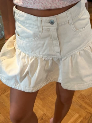 Vit jeansskort med volang - Superfin vit skort i jeansmaterial med volangkant och klassiska fickor fram. Hög midja och knappstängning framtill. Perfekt för en trendig och avslappnad look.