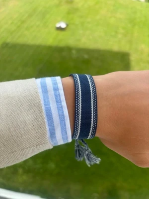 Marinblått tygarmband med tofsar - Snyggt marinblått tygarmband med vita detaljer och två dekorativa tofsar. Armbandet har ett brett band och knyts med flätade snören. Perfekt accessoar för en avslappnad och trendig stil.