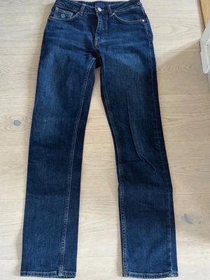 Mörkblå raka jeans - Säljer ett par mörkblå jeans med rak passform och klassisk femficksdesign. Jeansen har hög midja och stängs med knapp och dragkedja. Materialet är denim i bomull och de har kontrastsömmar. Perfekta för en clean och tidlös look.